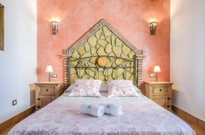 A bedroom with a stone-patterned headboard, two bedside tables with lamps, a bed with pink floral bedding, and two rolled towels on the bed.