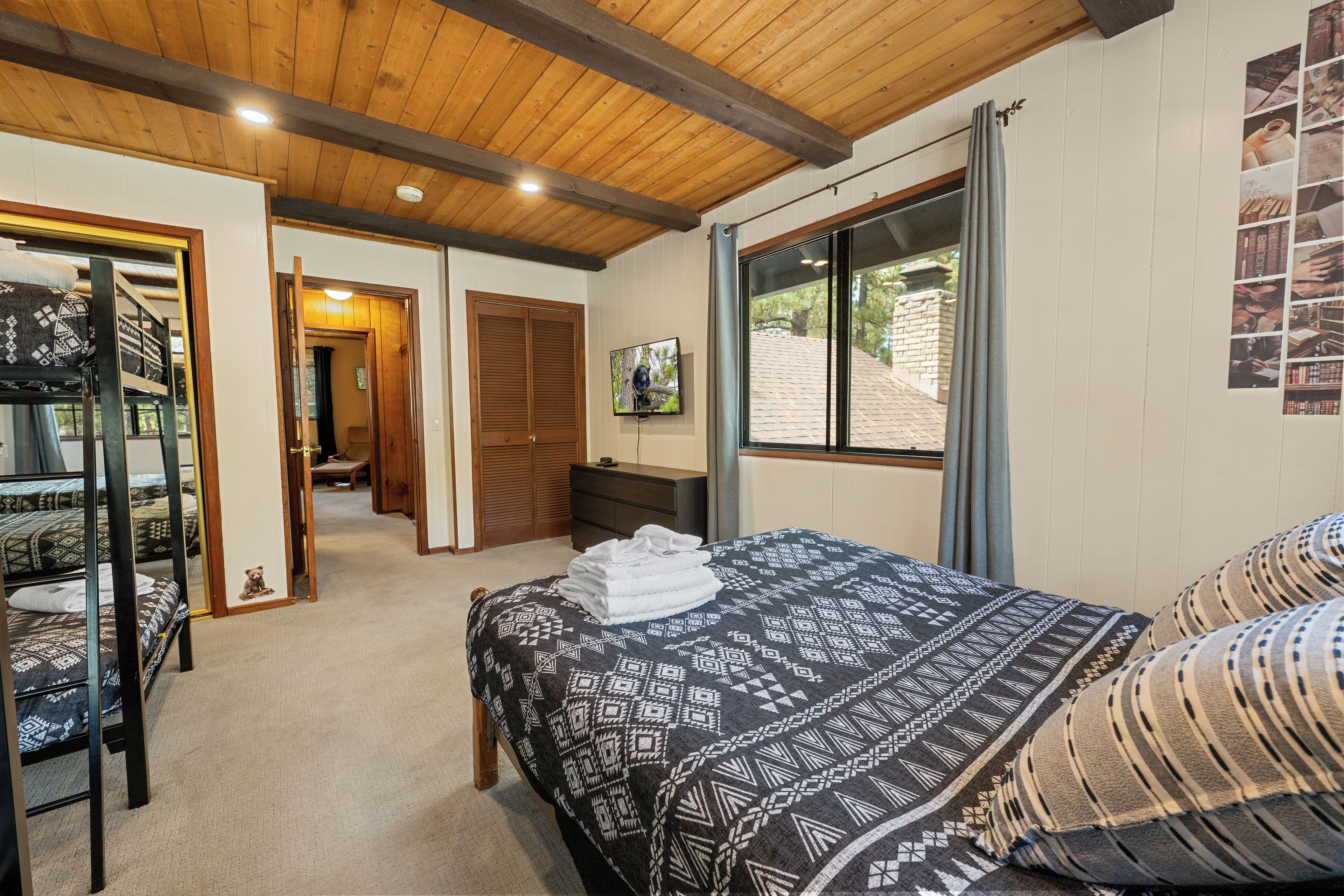 A bedroom with a bunk bed, a single bed with patterned bedding, a wooden ceiling, a window with curtains, and a wall-mounted picture.