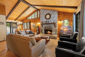 A cozy living room with a stone fireplace, wooden beams, a large sofa, and a television. - Bear Woods 3BR Retreat (Big Bear City)