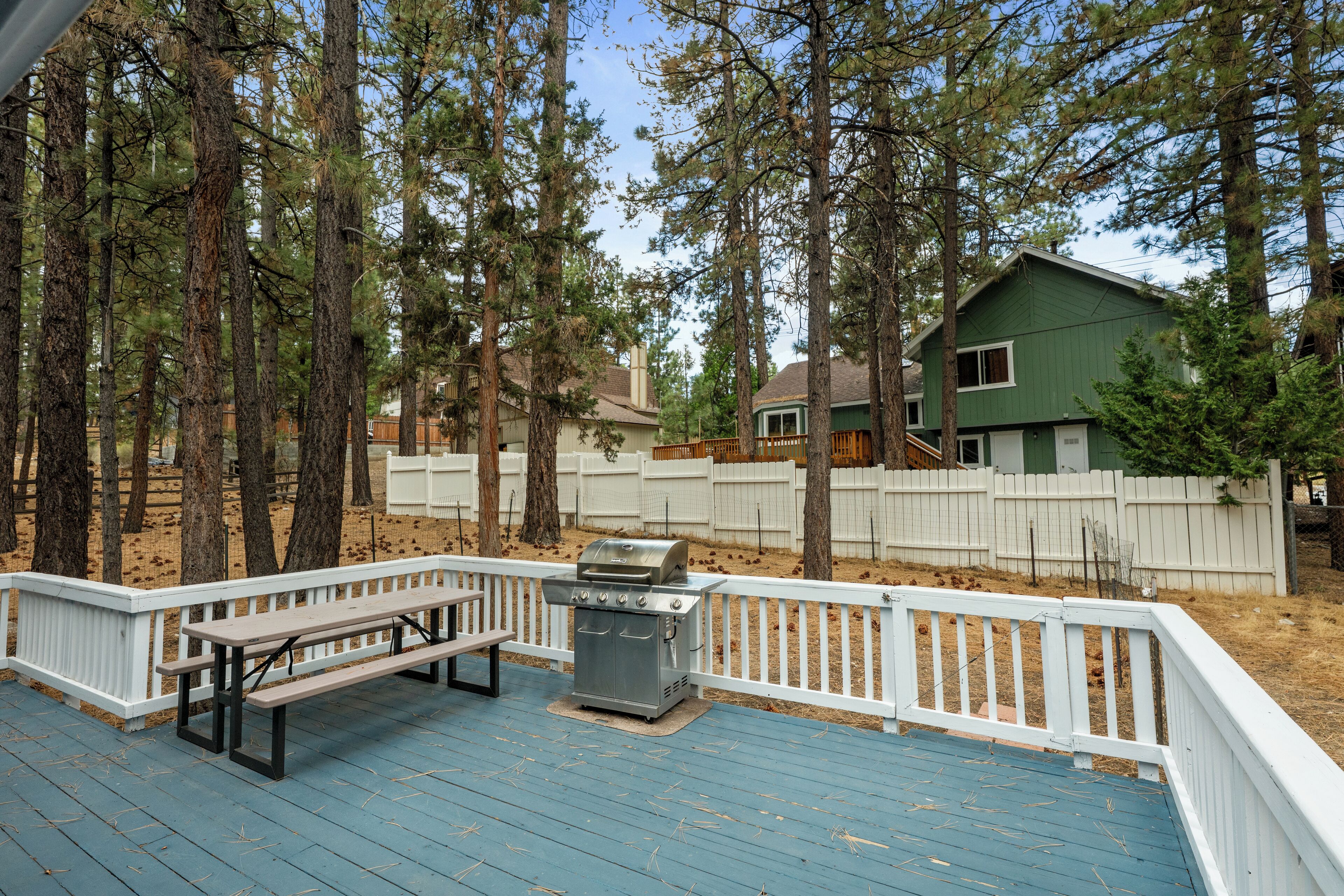 A deck with a grill and picnic table, surrounded by tall trees and a white fence.