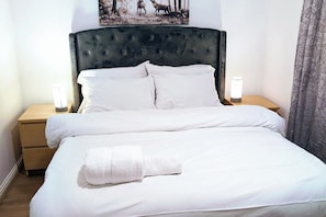 A neatly made bed with white linens and a folded towel, a tufted headboard, two bedside tables with lamps, and a floral curtain. - 2 Bed - Sleeps 4 - Free Parking - 5 Min to Centre (Manchester)