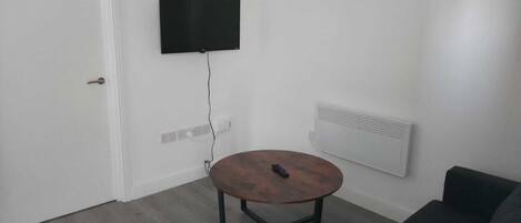 A small, sparsely furnished room with a round wooden table, a black couch, a mounted TV, and a white door.