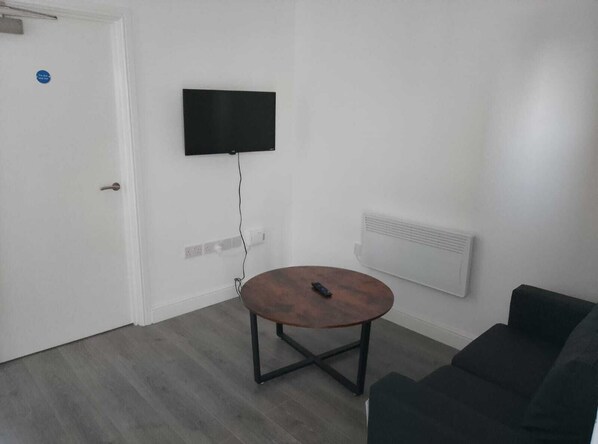 A small, sparsely furnished room with a round wooden table, a black couch, a mounted TV, and a white door. - Akai Manage (Scunthorpe)