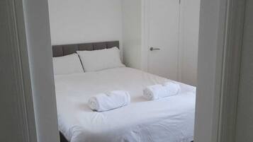 A neatly made bed with white linens and two rolled towels, a grey headboard, and a white door in a room with white walls and a grey carpet.