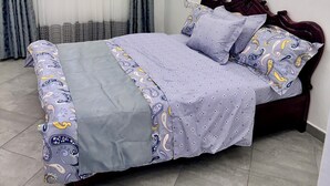 Uma cama bem arrumada com colcha estampada e almofadas, uma cabeceira de madeira escura e uma janela com cortinas finas.