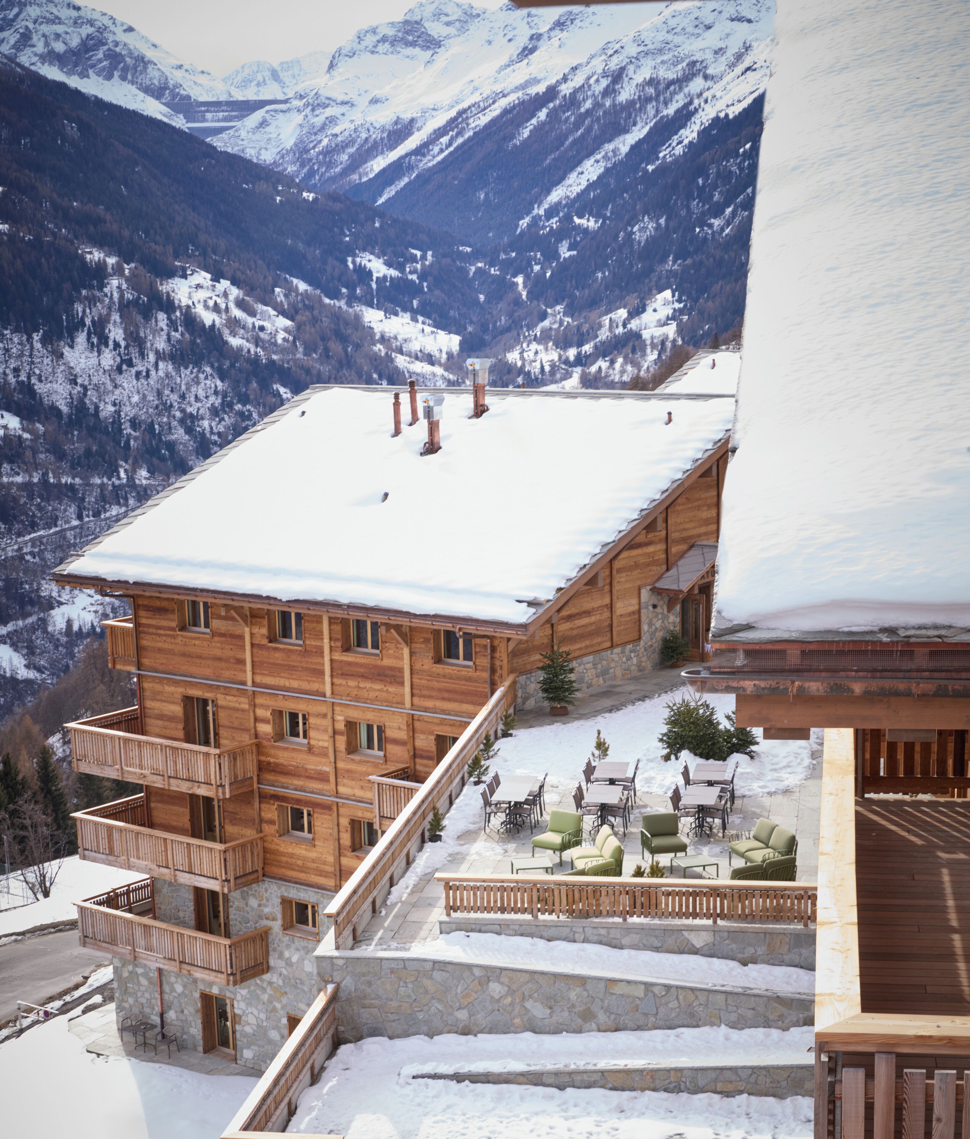 Un chalet en bois avec un toit enneigé, des murs en pierre et un balcon. Il dispose d’un espace extérieur aménagé et offre une vue sur des montagnes couvertes de neige.