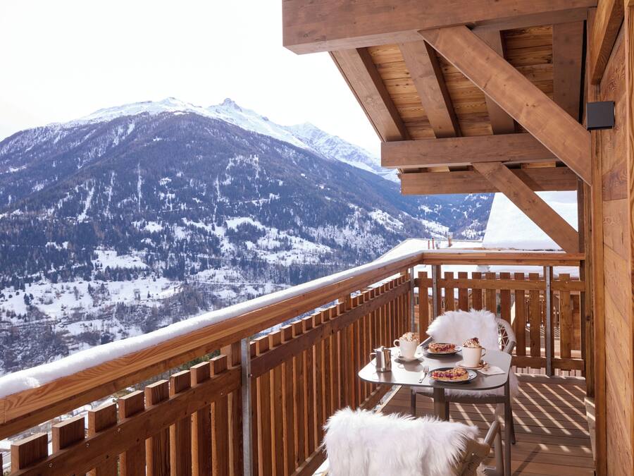 Un terrazzo in legno con tavolo e sedie, affacciato su una catena montuosa innevata.