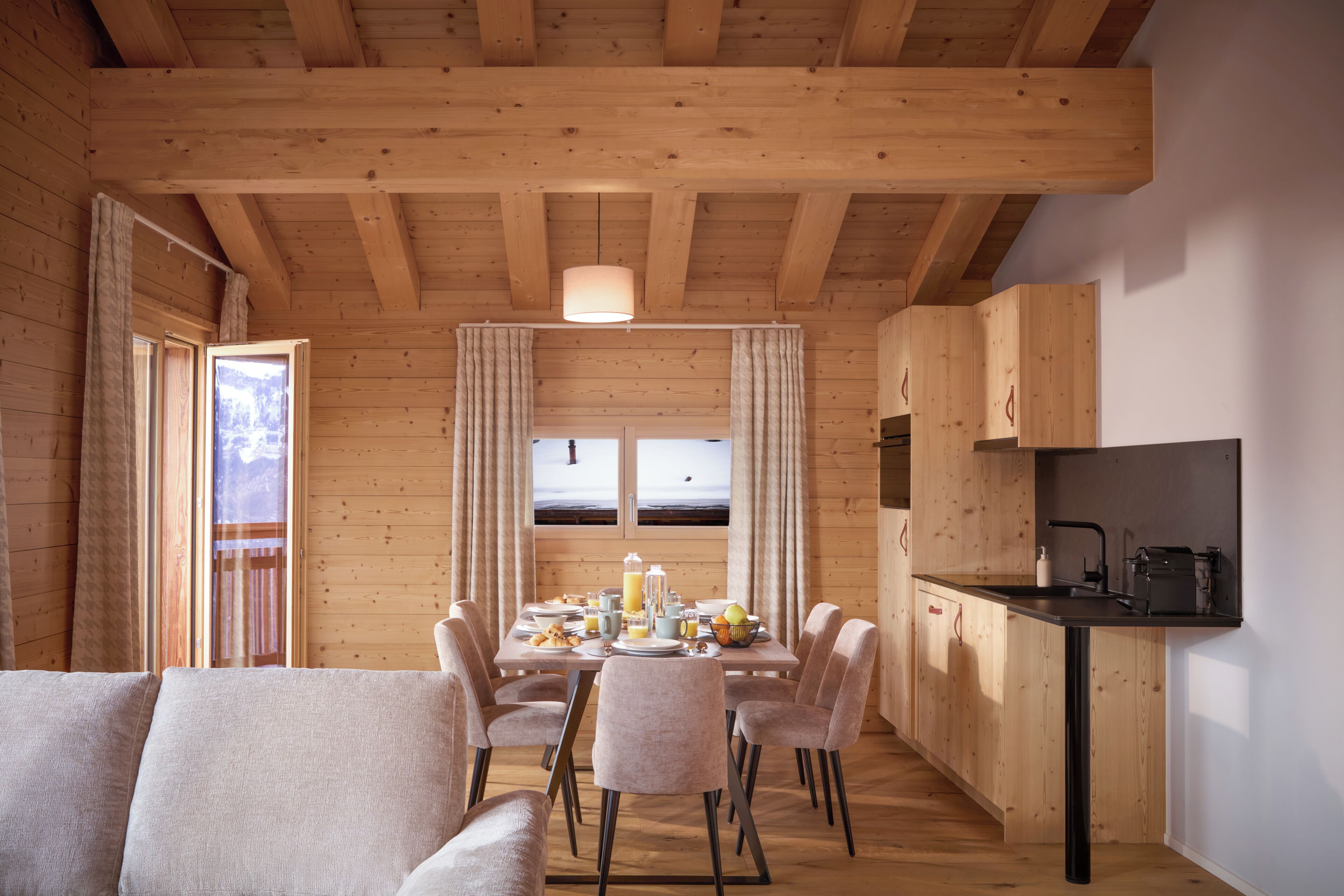 Een houten chalet van binnen met een eethoek, keuken en grote ramen.