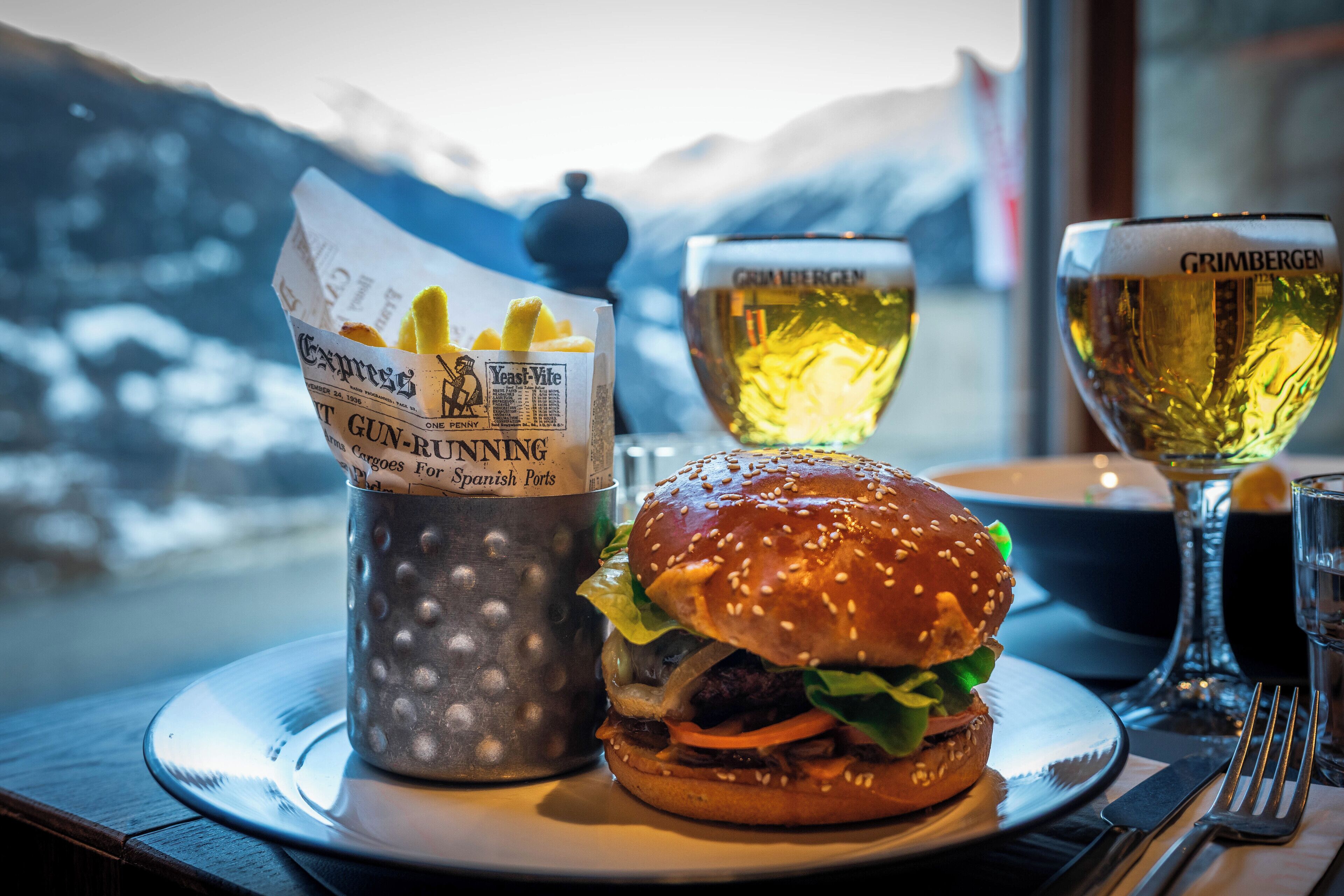 A meal with a burger and fries, a beer glass, and a menu on a table with a mountain view in the background.