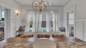 Una habitación amplia con una gran mesa de madera, un tapete tejido y una araña. Hay cortinas blancas y un banco con cojines de colores.