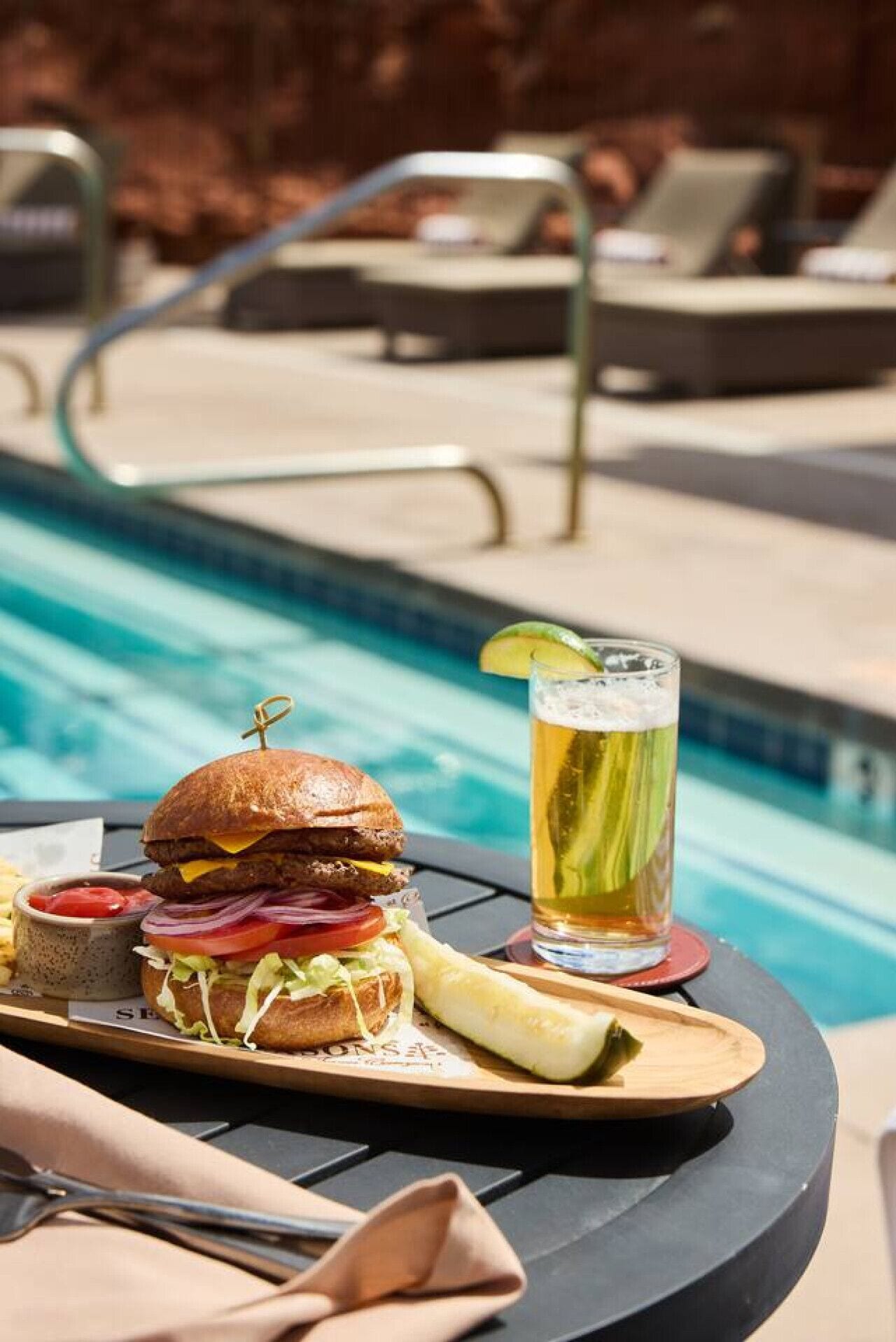 A poolside dining setup with a burger, fries, a pickle, and a beer.