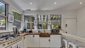 Une cuisine moderne avec des meubles blancs, une cuisinière à gaz, un four à micro-ondes et un évier. On y trouve trois tabourets et une fenêtre donnant sur des arbres.