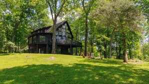 Une grande cabane à deux étages, entourée d’arbres, avec une pelouse verdoyante et un espace extérieur aménagé pour s’asseoir.