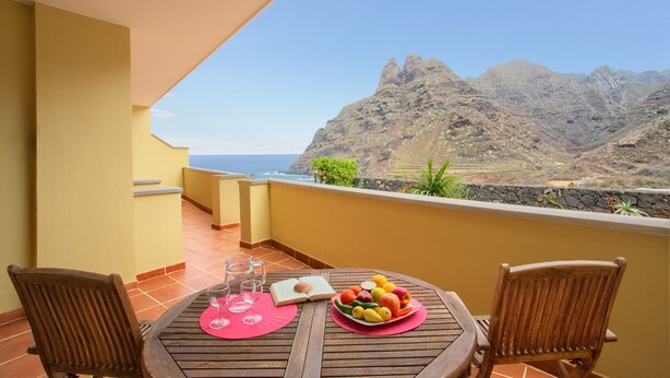 Ein Holztisch mit Obstschale und Buch, zwei Stühle und Blick auf Berge und Meer.