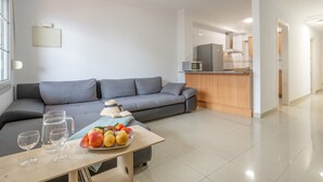 Ein modernes Wohnzimmer mit einem grauen Sofa, einem Couchtisch mit Früchten und Gläsern sowie einer Küche im Hintergrund.