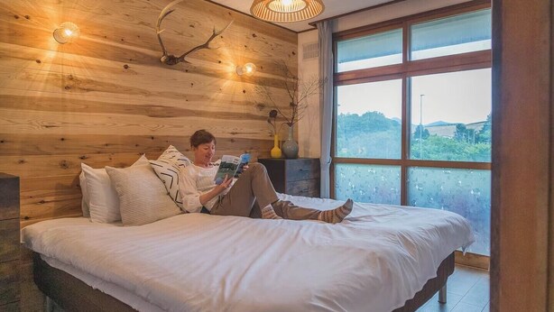 A bedroom with a wooden headboard, a large window with a view, and a person reading a book.