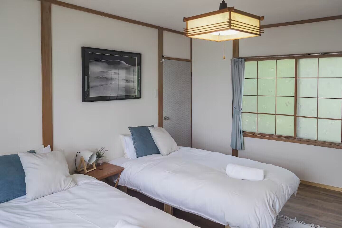 Ein Zimmer mit zwei Betten, einem gerahmten Bild an der Wand, einem Nachttisch mit Pflanze und einem Fenster mit Vorhängen.