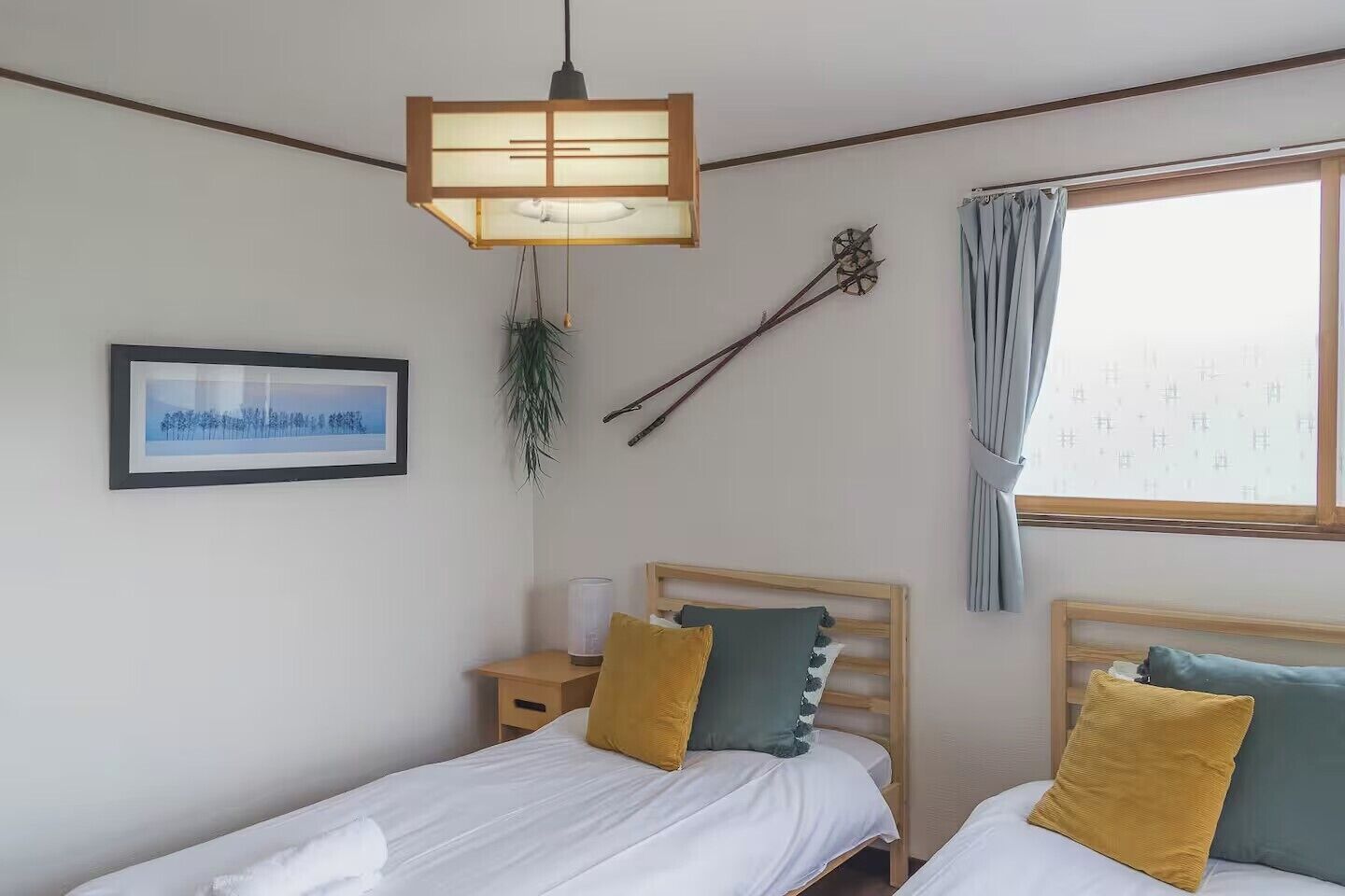 A bedroom with two beds, a bedside table, a hanging plant, a wall clock, and a window with curtains.