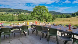 An outdoor dining area with a table set for six, surrounded by chairs, overlooking a scenic landscape with hills and trees.