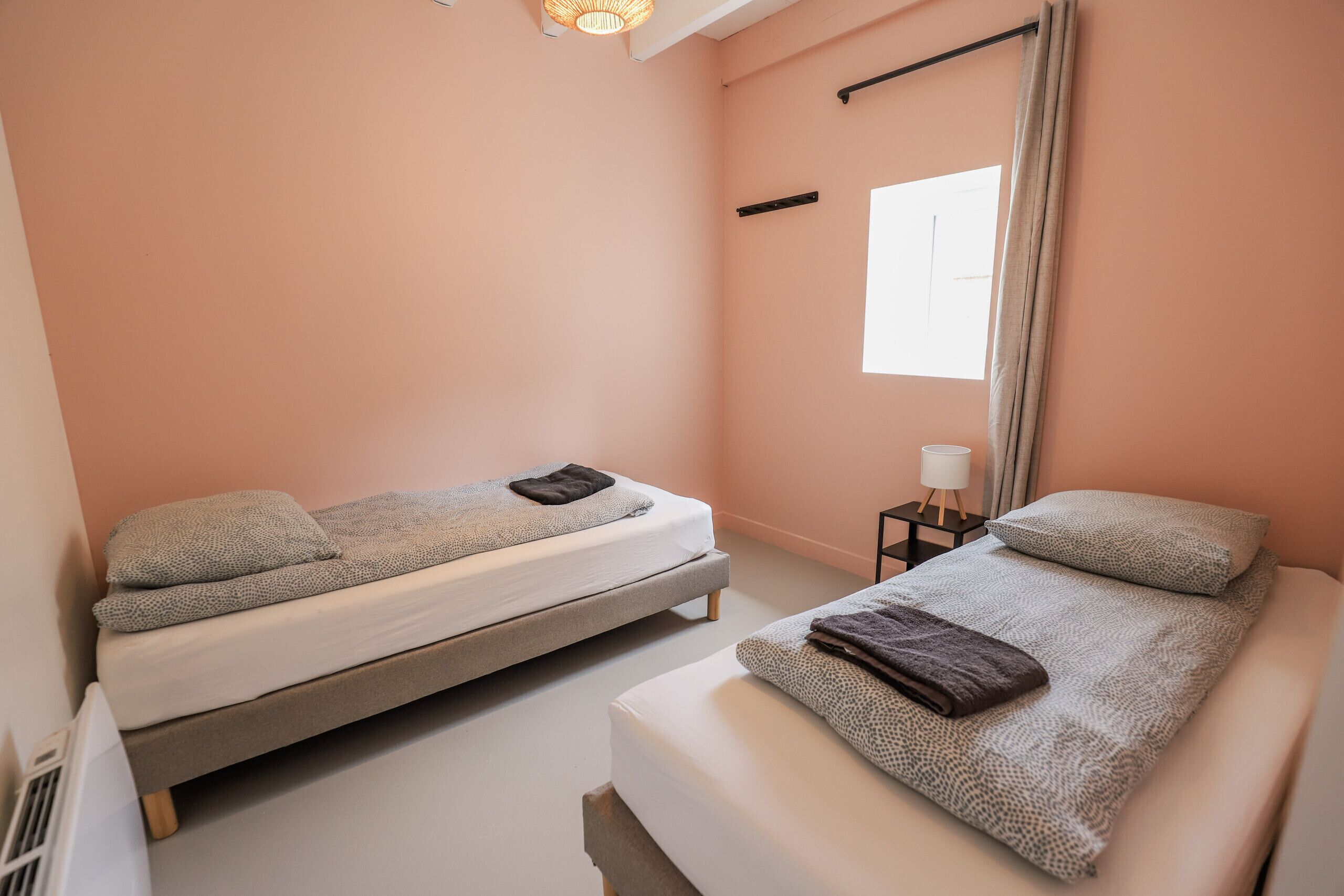 Une chambre avec deux lits simples, une table de chevet avec une lampe, une fenêtre avec des rideaux et un luminaire au plafond.