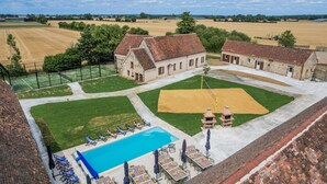 Una amplia piscina al aire libre con sillones y mesas, rodeada de un césped cuidado y un edificio de ladrillo.