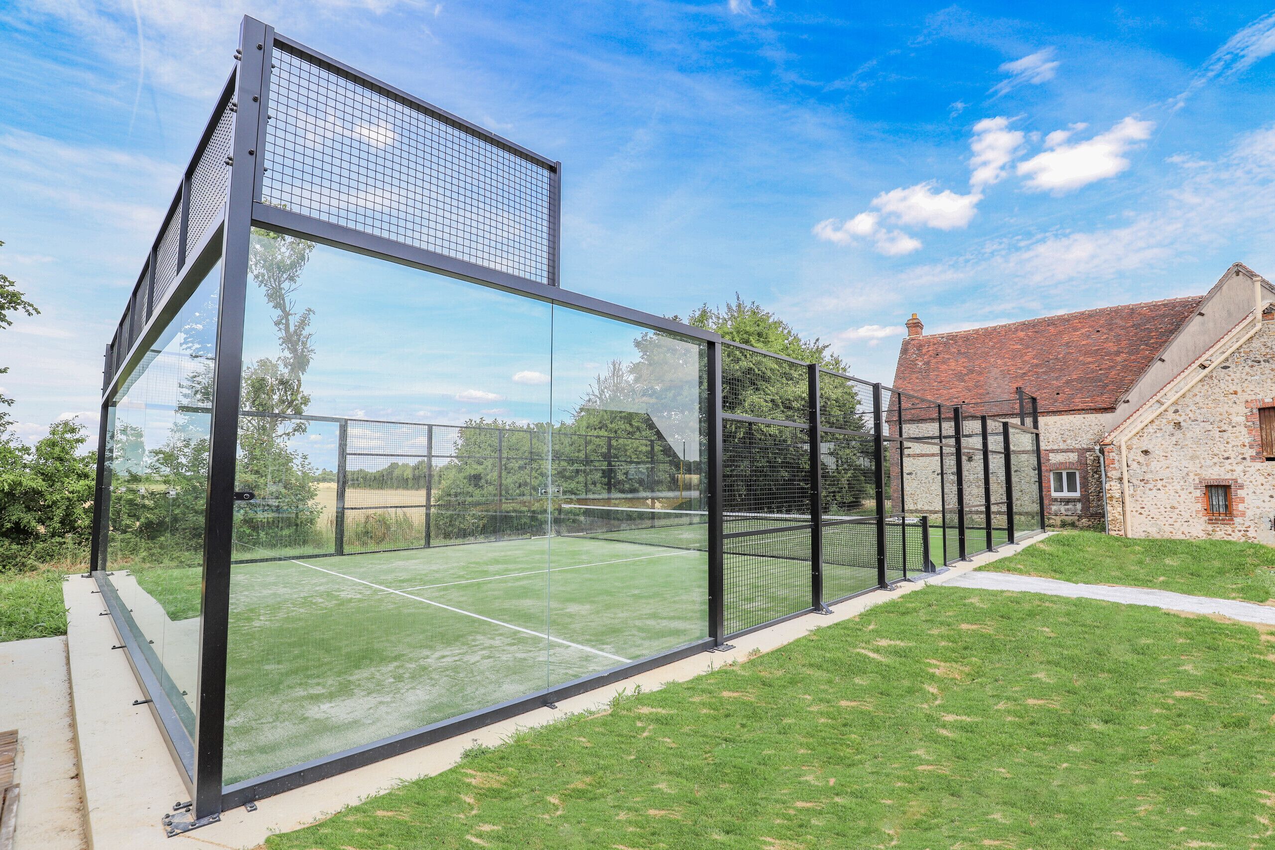 A tennis court enclosed by a glass fence with a stone building in the background.