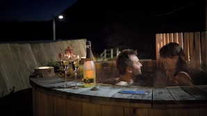 Un bain à remous où se trouvent deux personnes, une bouteille de champagne et deux verres de vin, le tout sur une terrasse en bois, la nuit.