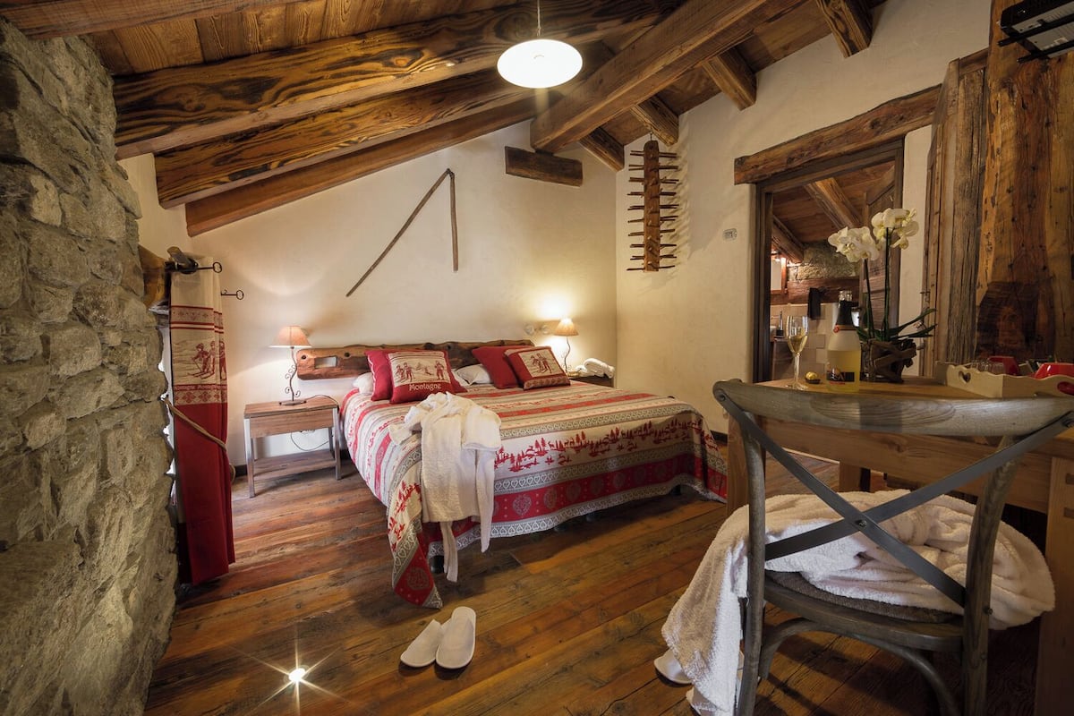 Ein rustikales Schlafzimmer mit Holzbalkendecke, einem Bett mit gemusterten Betttextilien, einem Nachttisch mit Lampe und einem Stuhl mit Handtuch.