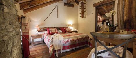 A rustic bedroom with a wooden ceiling, a bed with patterned bedding, a bedside table with a lamp, and a chair with a towel.