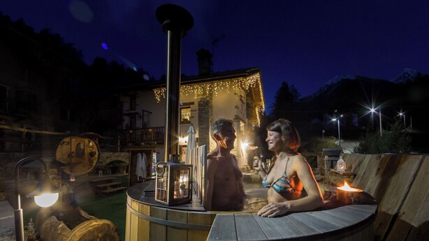 Un bain à remous en bois, avec une structure en pierre en arrière-plan, entouré d’un éclairage extérieur.