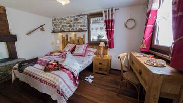 A cozy bedroom with a stone wall, a wooden bed, a desk with a chair, and a window with curtains.