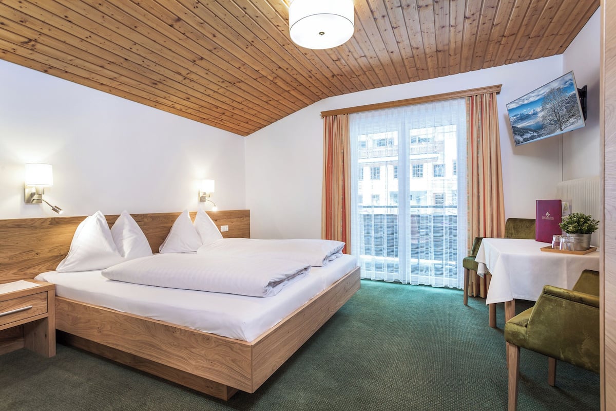 Habitación de hotel con techo de madera, una cama grande con ropa blanca, un escritorio con silla y un televisor montado en la pared.