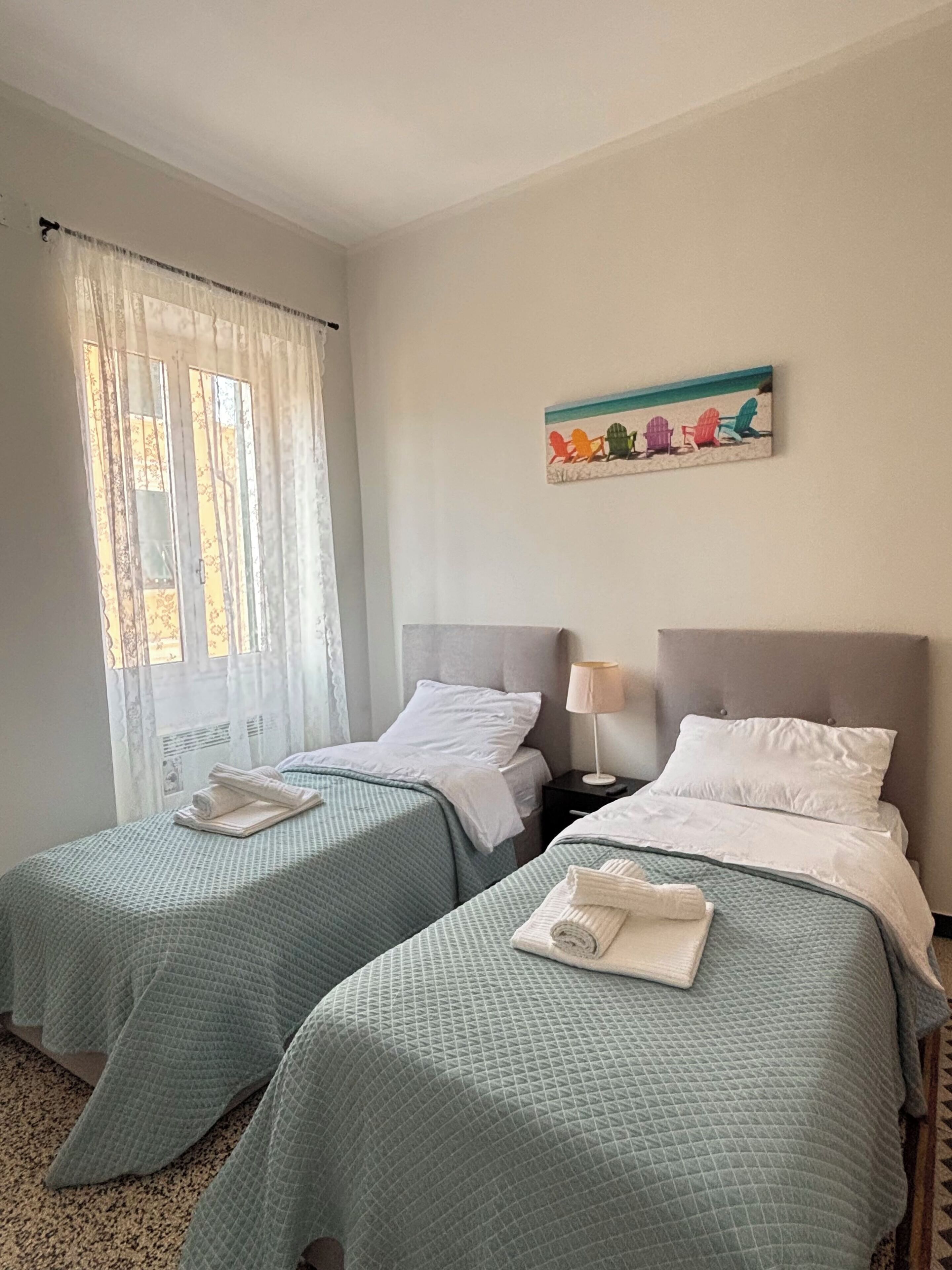 Een hotelkamer met twee bedden, elk voorzien van een groene geborduurde sprei en witte kussens. Aan de muur hangt een schilderij van strandstoelen, en er is een raam met dunne gordijnen.