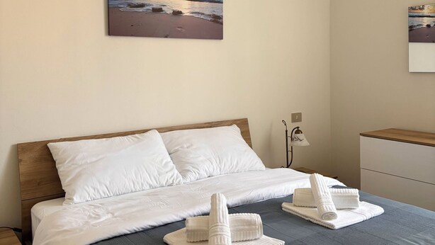 A neatly made bed with white bedding and two rolled towels, a painting of a sunset over the ocean on the wall, and a bedside lamp. - Three-room apartment just a few steps from the beaches in the center of Imperia (Imperia)