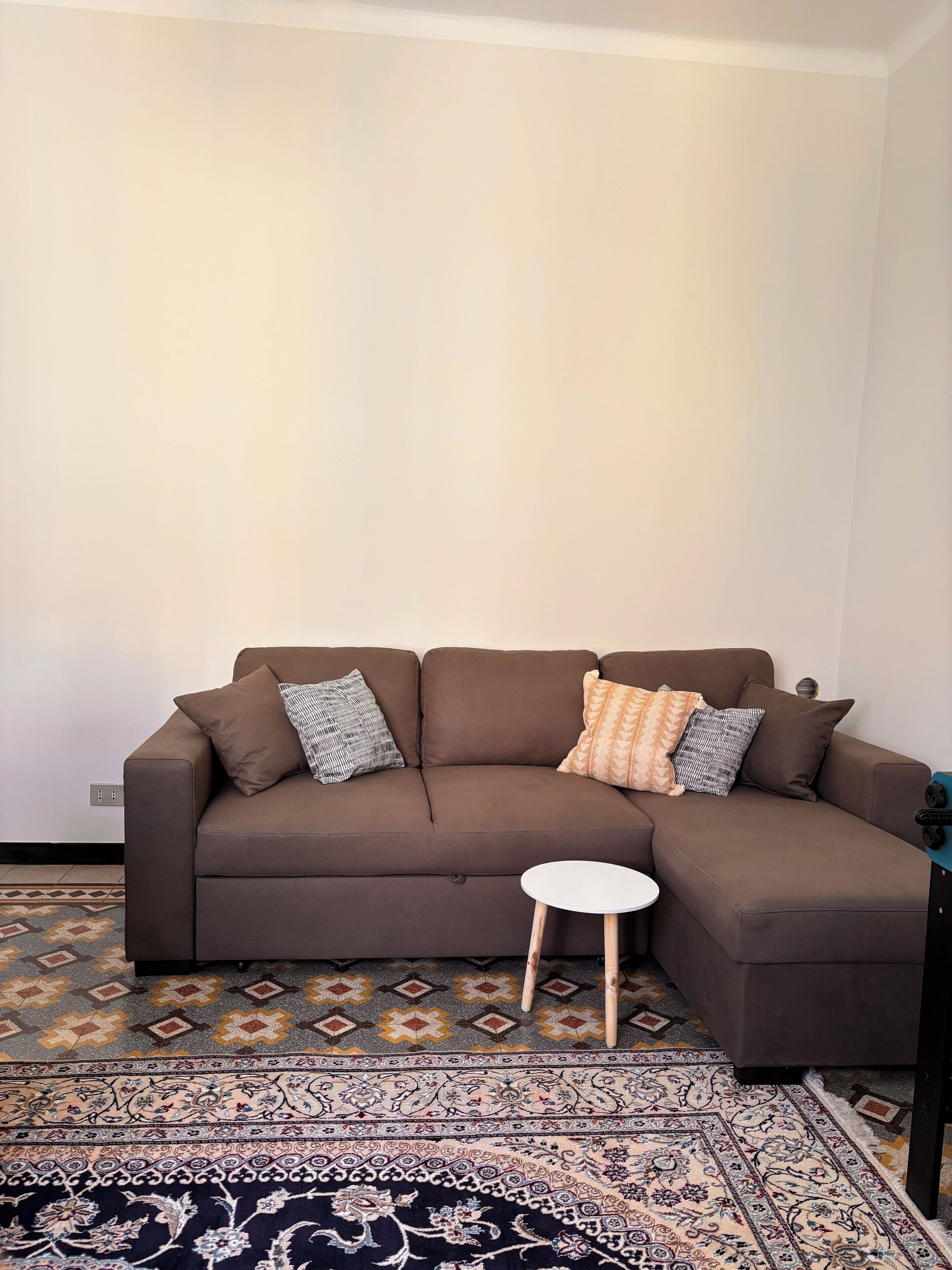 A brown sectional sofa with patterned cushions, a small round side table, and a patterned rug in a room with plain walls and a patterned floor.