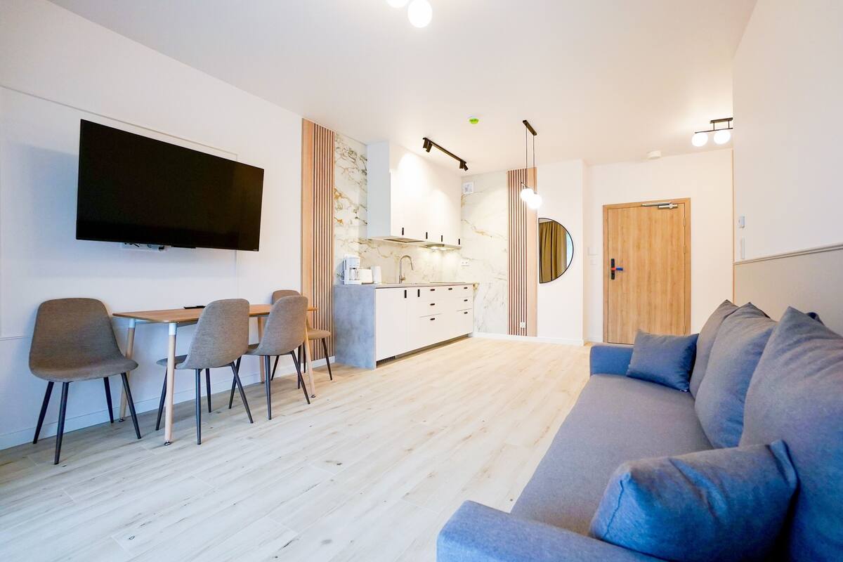 Un salon moderne avec un canapé gris, un coin repas avec une table et des chaises, une cuisine avec un dosseret en marbre et un téléviseur fixé au mur.