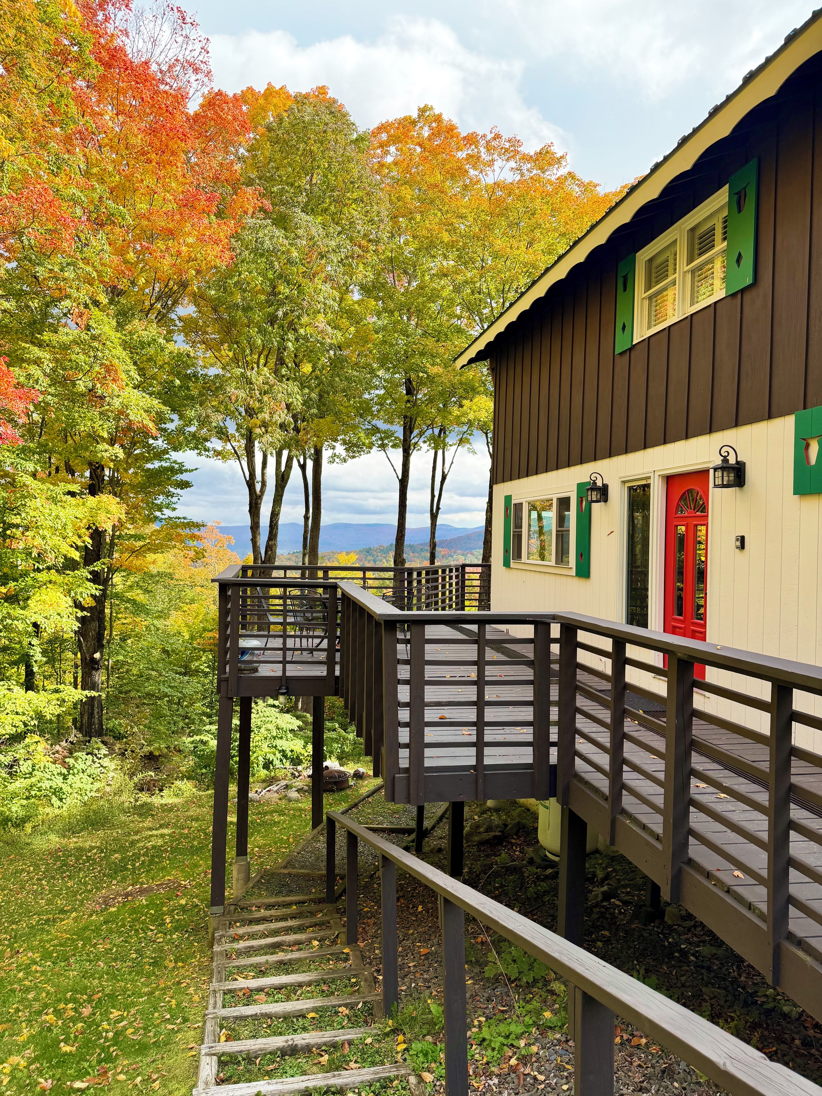 A wooden deck with a staircase leading to a building with a red door and green shutters.