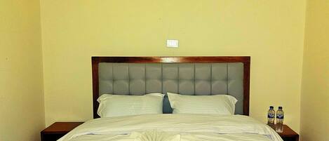 A neatly made bed with a grey headboard, two bedside tables with water bottles, and a plain wall.