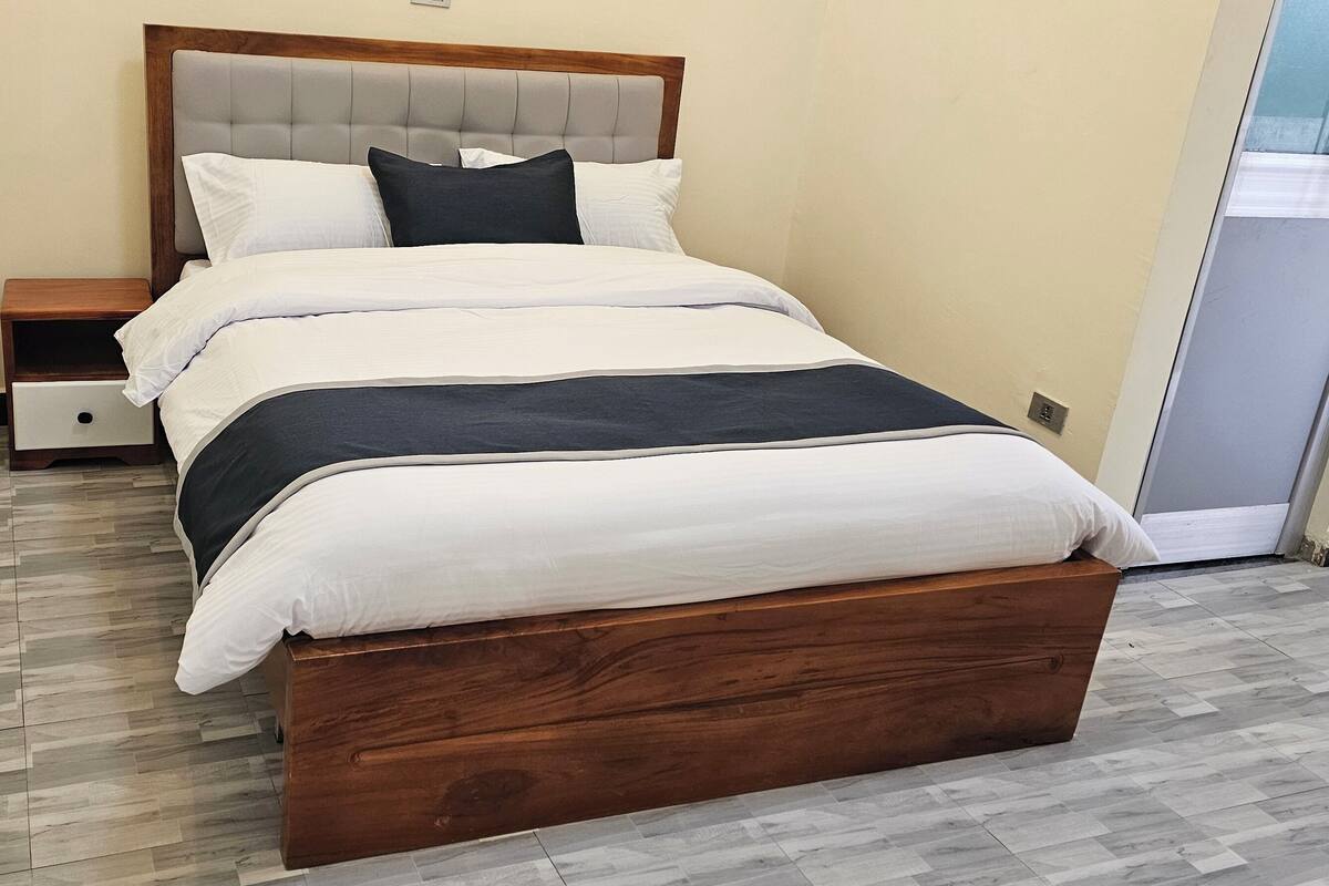 A neatly made bed with a wooden headboard, a bedside table, and a small drawer unit.