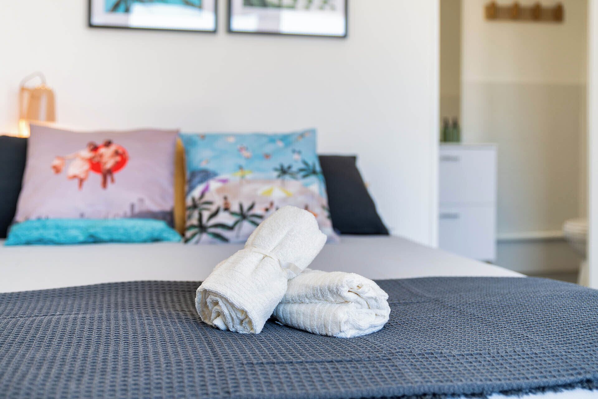 A neatly made bed with a rolled white towel, a gray textured blanket, and two decorative pillows with floral designs.