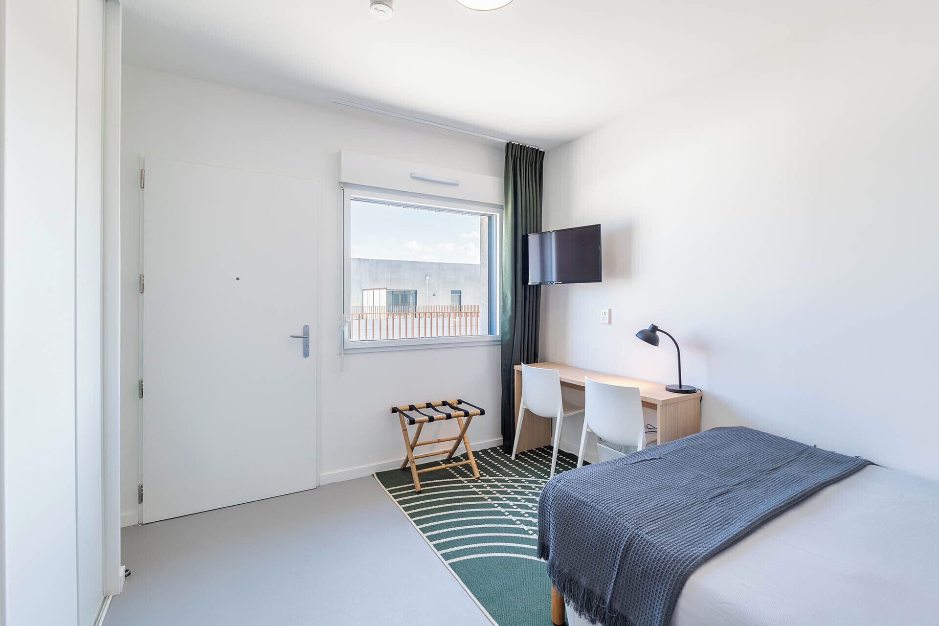 Une chambre d’hôtel de taille réduite, comprenant un lit, un bureau, une chaise, un téléviseur et une fenêtre donnant sur un immeuble.