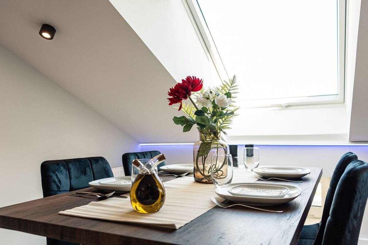 A dining table set with plates, cutlery, and a bottle of olive oil, with a vase of flowers and a glass of water on the table.