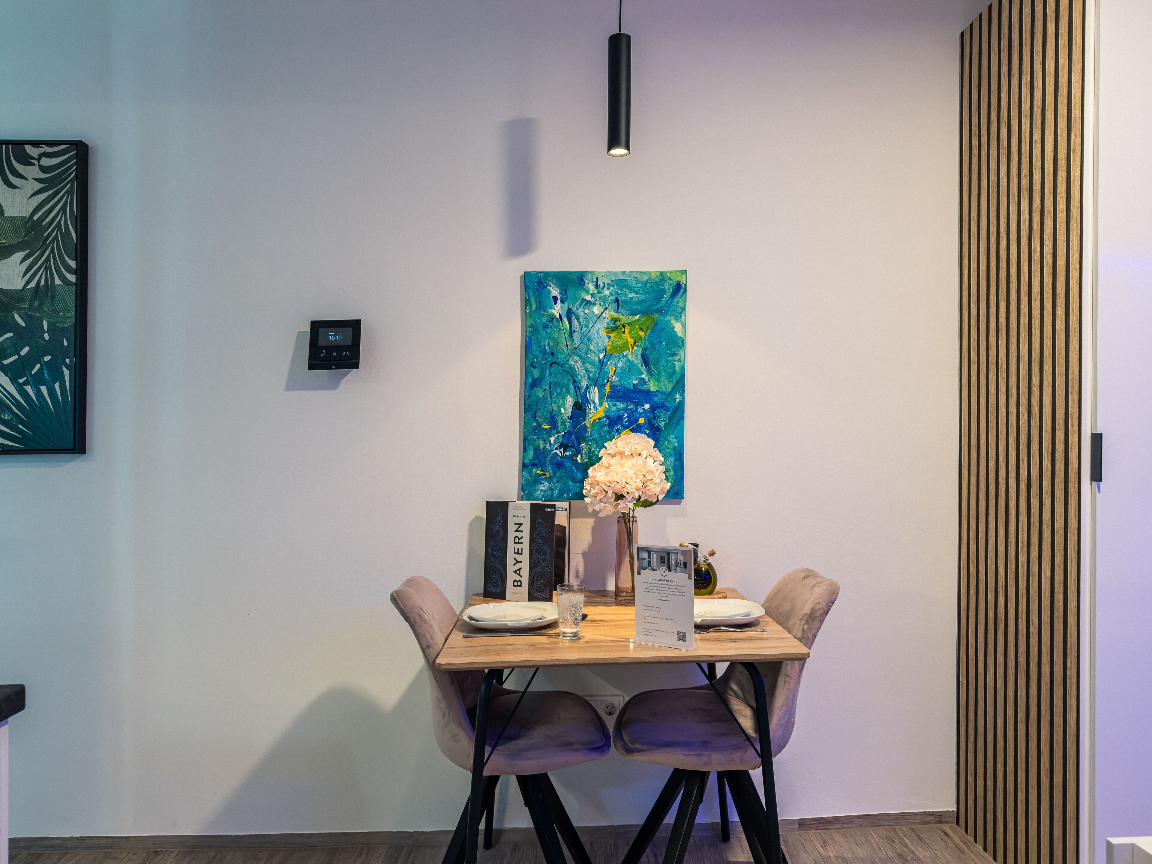 A dining area with a wooden table, two chairs, a vase with flowers, a painting, and a thermostat on the wall.