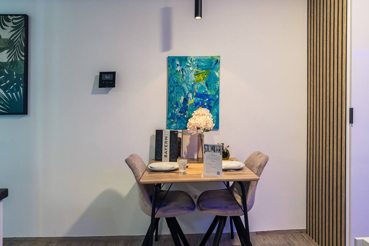 A dining area with a wooden table, two chairs, a vase with flowers, a painting, and a thermostat on the wall.