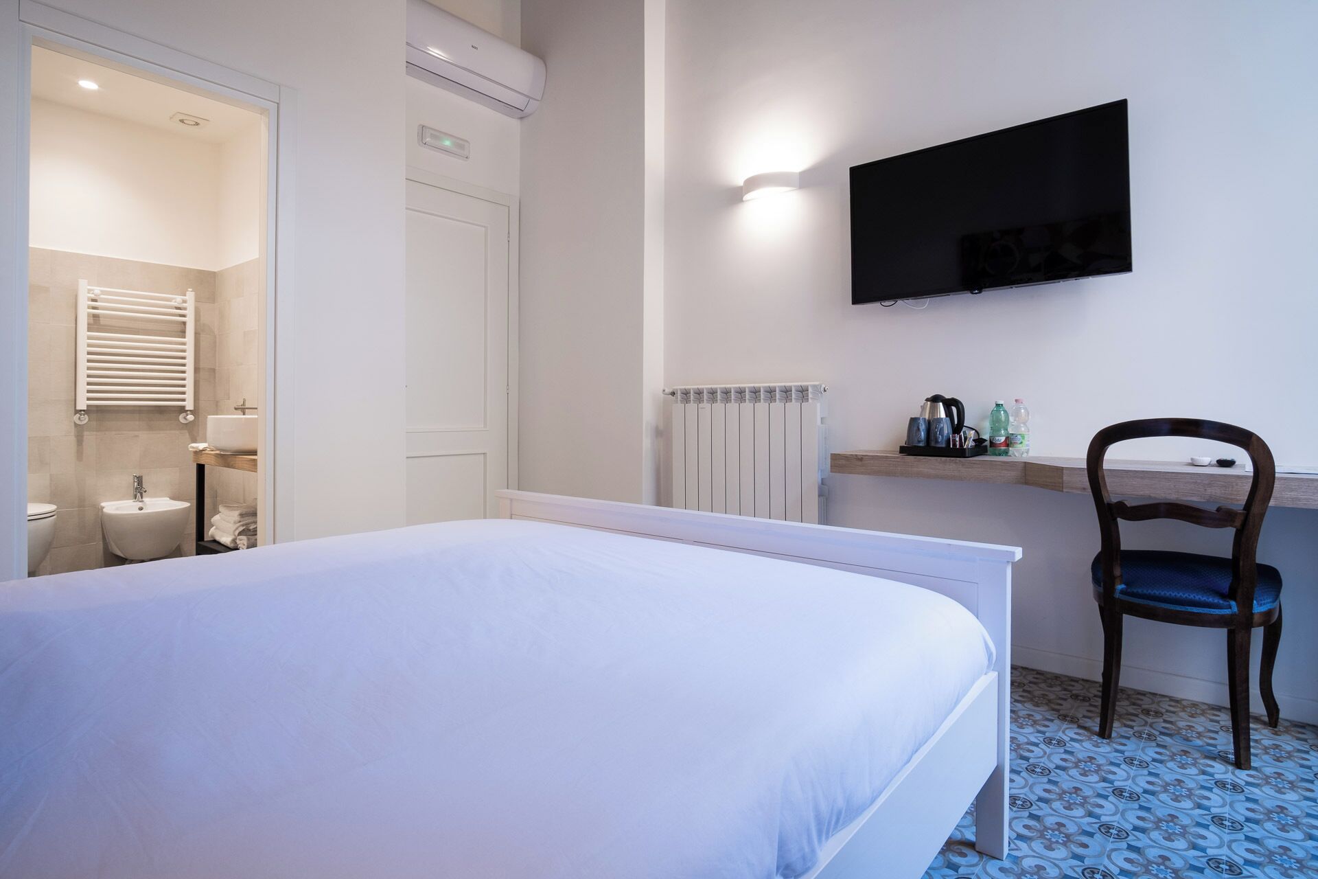 A bedroom with a bed, a desk with a chair, a TV mounted on the wall, and a radiator.