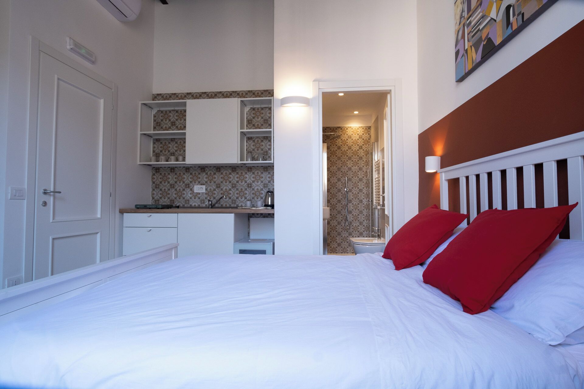 A bedroom with a bed, red pillows, a white headboard, a bathroom with a shower, and a kitchen area with cabinets and a countertop.