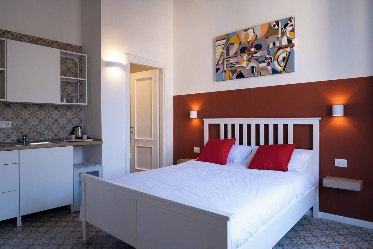 Un dormitorio con una cama blanca, almohadas rojas y blancas, un cabecero blanco y un cuadro abstracto colgado en la pared.
