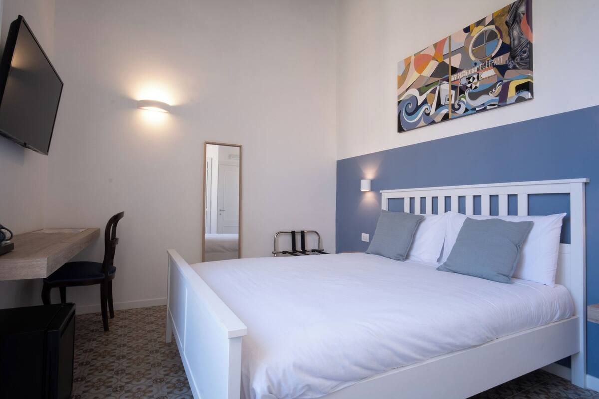 Un dormitorio con una cama, un escritorio, una silla, un televisor y una obra de arte en la pared.