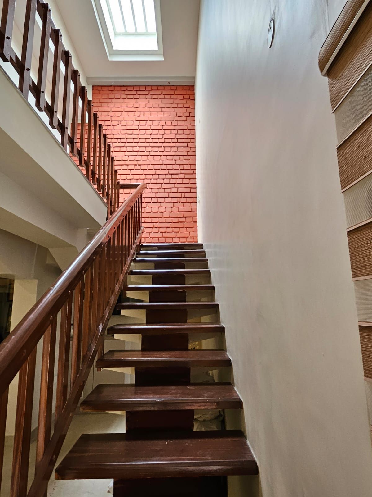 A staircase with wooden handrails and steps, leading to a room with a red brick wall and a skylight.