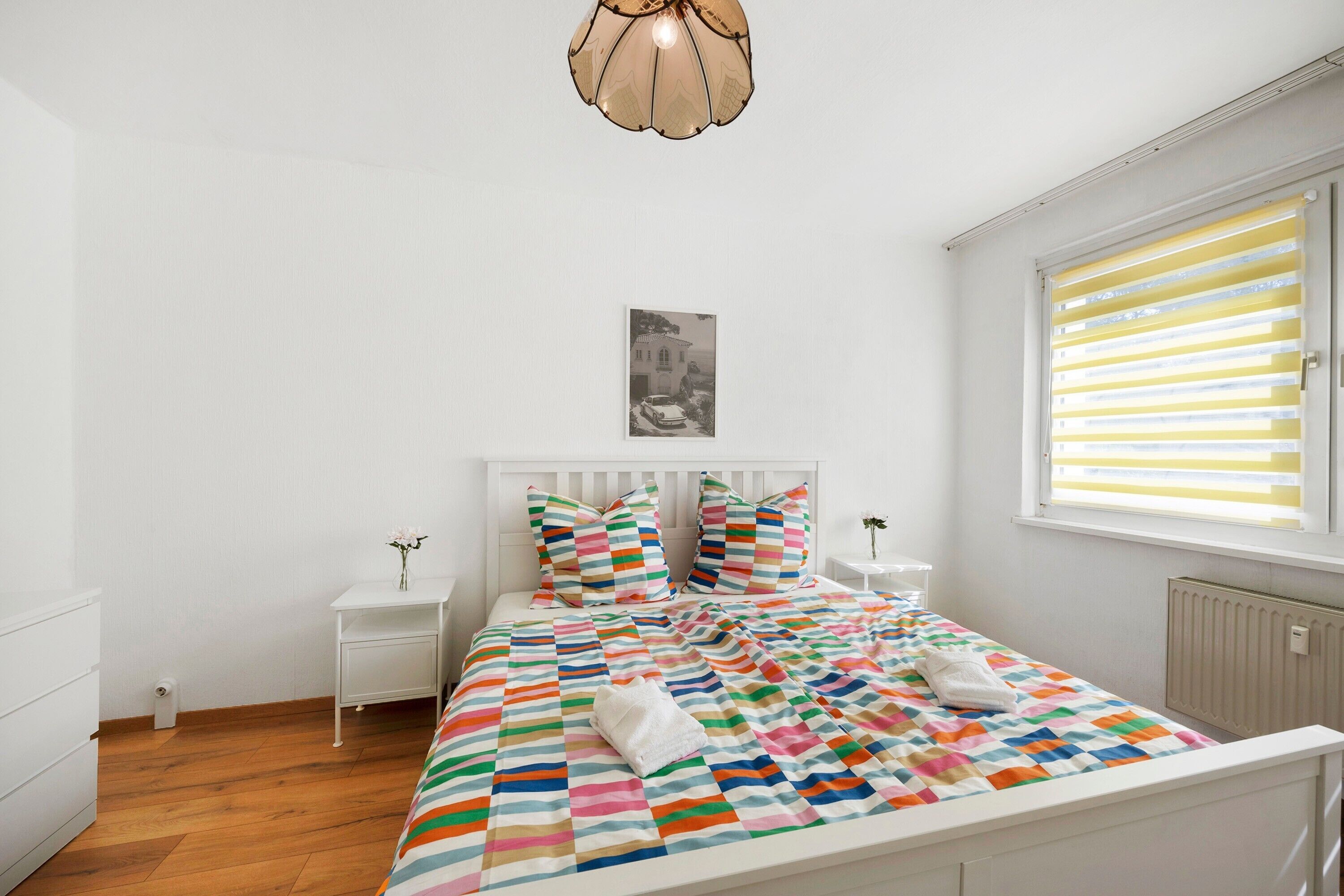 Une chambre à coucher avec un lit, une tête de lit blanche, des tables de chevet, une fenêtre avec des stores et un tableau accroché au mur.